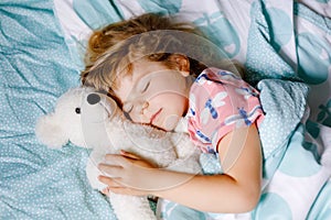 Cute little toddler girl sleeping in bed with favourite soft plush toy teddy bear. Adorable baby child dreaming, healthy