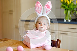 Cute little toddler girl wearing Easter bunny ears playing with colored pastel eggs. Happy baby child unpacking gifts