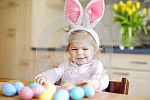 Cute little toddler girl wearing Easter bunny ears playing with colored pastel eggs. Happy baby child unpacking gifts