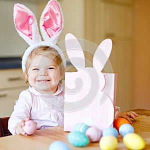 Cute little toddler girl wearing Easter bunny ears playing with colored pastel eggs. Happy baby child unpacking gifts