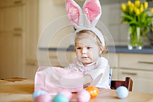 Cute little toddler girl wearing Easter bunny ears playing with colored pastel eggs. Happy baby child unpacking gifts