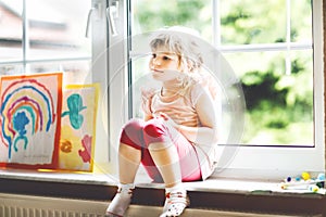 Cute little toddler girl by window with rainbow painted with colorful colors. Preschool on bad weather rainy day looking