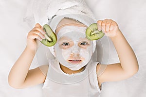 Cute little two three years old girl with a towel over her wet hair, facial mask on her face, relaxed and lying on white