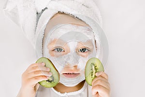 Cute little two three years old girl with a towel over her wet hair, facial mask on her face, relaxed and lying on white