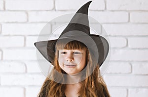 Cute Little Witch In A black Hat Against A white brick wall.Portrait of a 4-year-old girl at the Halloween festival.A pretty