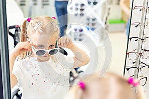 Cute little young caucasian blond girl trying on and choosing sunglasses in front of mirror at optic eyewear store. Adorable