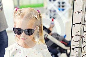 Cute little young caucasian blond girl trying on and choosing sunglasses in front of mirror at optic eyewear store. Adorable