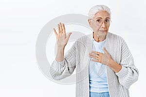 Cute lively old woman in glasses, grandmother look camera sincere, press hand to heart and raise arm to swear shes