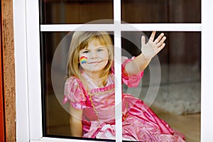 Cute lonely little toddler girl in princess dress sitting by window with rainbow with colorful colors on face during