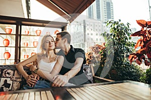 Cute loving couple sitting in cozy restaurant and terrace on sunset background and waiting for order. Young man hugs and kisses a