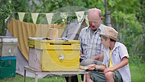 cute male child helping his grandfather beekeeper paint beehive with yellow paint, smile, look at camera and point up