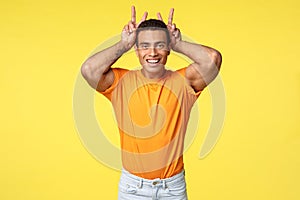 Cute masculine and silly hispanic boyfriend in orange t-shirt, make peace gesture, bunny ears behind head, smiling