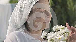 Cute mature woman with white shawl on her head tears off daisy petals at the clothesline outdoors. Washday. Positive