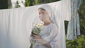 Cute mature woman with white shawl on her head tears off daisy petals at the clothesline outdoors. Washday. Positive