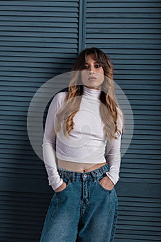 Cute model of a modern young woman in a white vintage top in jeans trendy pants with red curly hair posing near a stylish wooden