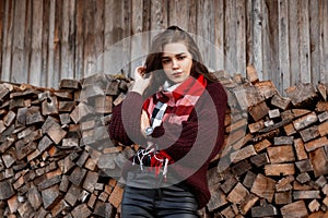 Cute model of a rural young woman in a stylish knitted sweater with a fashionable scarf stands near an old wooden wall with logs.