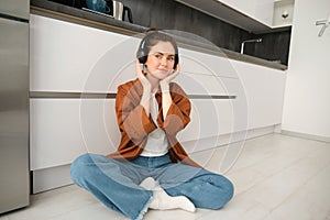 Cute modern girl on kitchen floor at home, listening music in wireless headphones, having fun