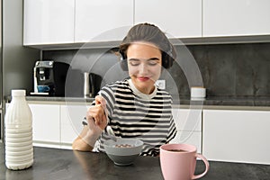 Cute modern woman, student eating quick breakfast, having cereals with milk and coffee, listening to podcast or music in