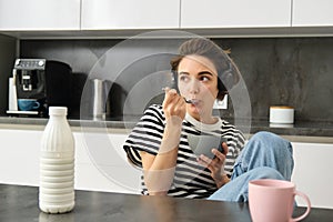 Cute modern woman, student eating quick breakfast, having cereals with milk and coffee, listening to podcast or music in