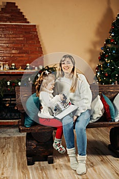 Cute mom and daughter are looking through a family photo album on Christmas Eve, spending time together by the fireplace