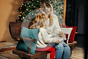 Cute mom and daughter are looking through a family photo album on Christmas Eve, spending time together by the fireplace