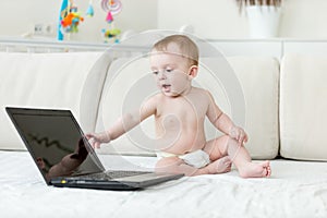 Cute 10 months old baby boy in diapers sitting on bed and looking at laptop