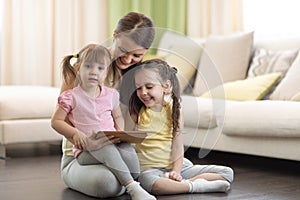 Cute mother with her 2 years old little daughter and 5 years old daughter playing with digital tablet at home