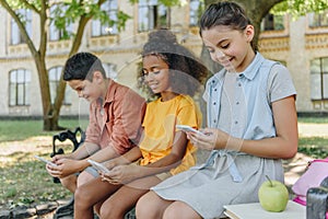 Cute multicultural schoolkids using smartphones while sitting on bench in schoolyard