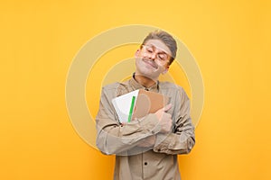 Cute nerd in glasses and mustache with books and exercise books in hands isolated on yellow background, smiling with closed eyes.