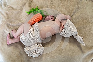 cute newborn baby with a carrot plush toy sleeping on a beige blanket, infant wears knitted rabbit costume