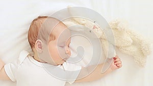 cute newborn baby girl is sleeping sweetly with a plush bunny in her hands on a white cotton bed at home, a quiet baby