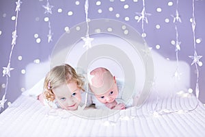 Cute newborn baby and his beautiful toddler sister playing together in a white bed between purple lights