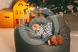 A cute newborn baby in a plaid jacket and a knitted hat is sleeping on a pouf in the background of a Christmas tree with