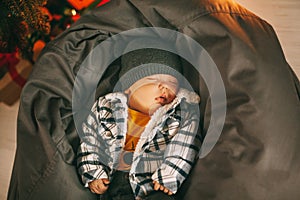 A cute newborn baby in a plaid jacket and a knitted hat is sleeping on a pouf next to a Christmas tree. new years eve