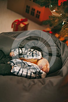 A cute newborn baby in a plaid jacket and a knitted hat is sleeping on a pouf next to a Christmas tree. new years eve