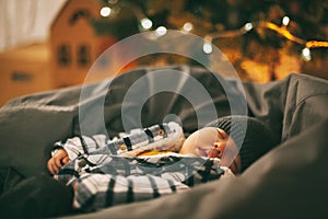 A cute newborn baby in a plaid jacket and a knitted hat is sleeping on a pouf next to a Christmas tree. new years eve