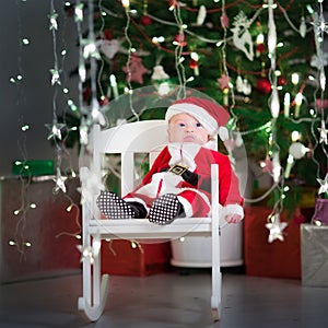 Cute newborn baby in a santa costume and hat sitting under Christmas tree