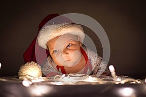 Cute newborn baby with santa hat raised his head over the lights under christmas tree