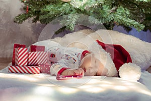 Cute newborn baby sleeping under Christmas tree near red gifts wearing Santa Claus hat