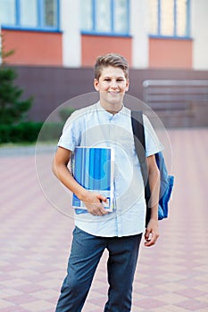 Cute, nice, young 11 years old boy in blue shirt stands with workbooks and backpack in front of his school. Education