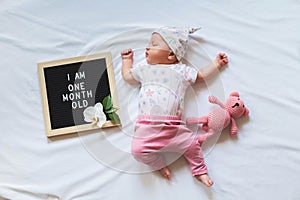 Cute one month old baby girl in trendy outfit laying between letter board and teddy bear. One month announcement.