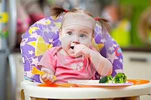 Cute one year old baby girl in highchair with pasta in kitchen at home