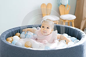 A cute one-year-old baby in a pink dress is bathing in a toy pool with colored balls and smiling