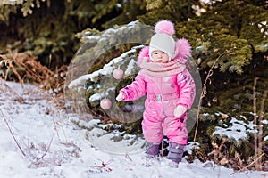 Cute one-year-old girl in a pink winter jumpsuit and boots stands in winter near the Christmas tree