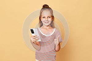 Cute optimistic smiling little brown haired girl wearing striped t-shirt standing isolated over beige background holding