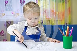 Cute pensive caucasian boy 3 years old drawing with colored pencil in a notebook sitting at a desk in a classroom in a kindergarte