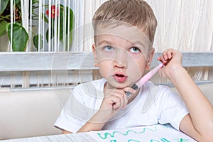 A cute pensive little boy holds a felt-tip pen in his hands and thinks over a drawing for mom. Drawing training