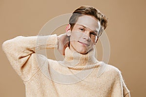 a cute, pleasant young man in a beautiful beige sweater is standing looking at the camera with his hand behind his head