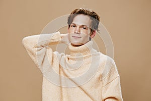 a cute, pleasant young man in a beautiful beige sweater is standing looking at the camera with his hand behind his head
