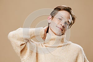 a cute, pleasant young man in a beautiful beige sweater is standing looking at the camera with his hand behind his head